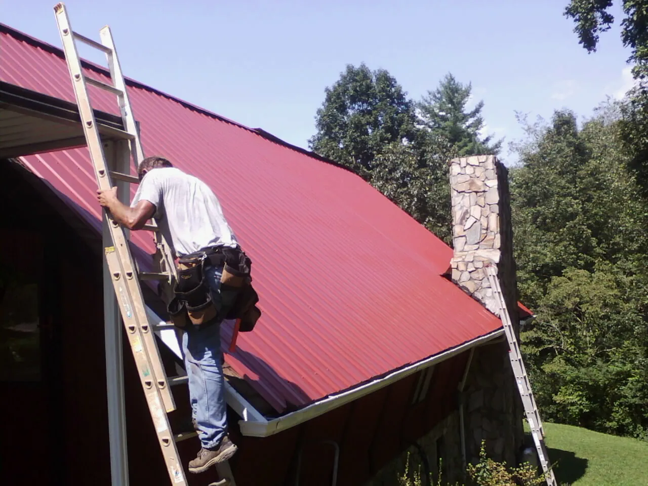 Roofer on a ladder beside a red metal roof and stone chimney — Soffit & Fascia Repair by Roofing Brothers in York Beach